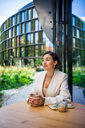 Young business woman holding and drinking coffee in street cafe in city. Portrait of beautiful woman worker waiting client on street of Pragueの写真素材