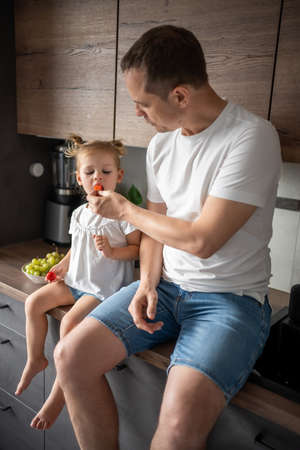 Cute little girl and her handsome dad are eating fruit in modern kitchen. Healthy eating.の写真素材