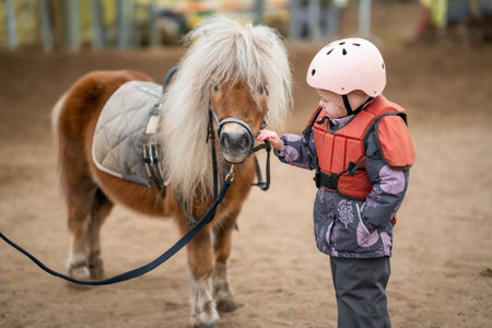 Portrait of little girl in protective jacket and helmet with her brown pony before riding Lesson. High quality photoの写真素材