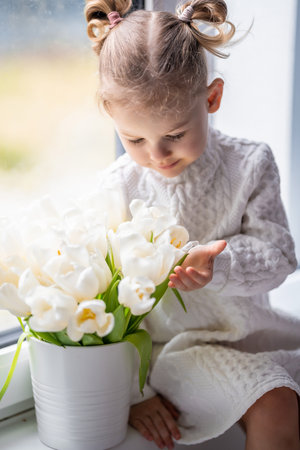 Little girl sitting by window with tulip flowers bouquet. Happy child, indoors. Mothers day, valentines day or birthday concept. High quality photoの写真素材