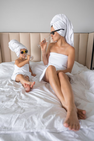 Happy family mother and little daughter in dressing gowns and towels laugh and drinking water with glasses during spa day at home. High quality photoの写真素材