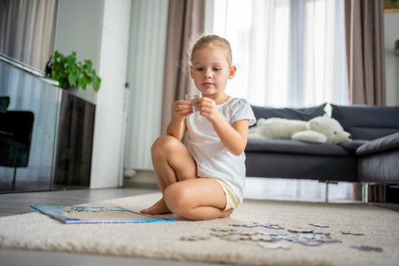 Little blonde girl sits at home on the carpet and collects puzzles. High quality photoの写真素材