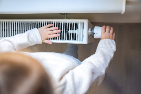 Little girl in warm sweater and socks standing by window turning the battery heating knob and warming up from the heating radiator. Heating in an apartment, at home. High quality photoの写真素材