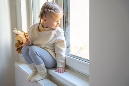 Little girl in warm sweater and socks sitting and looking out the window, holding autumn leaf and warming up from the heating radiator. Heating in an apartment, at home. High quality photoの写真素材