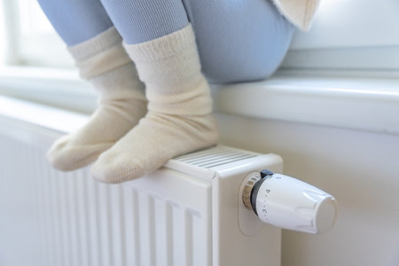 Feet of little girl in warm white socks. Child sitting and warming up from the heating radiator. Focus on battery heating knob. Heating in an apartment, at home. High quality photoの写真素材