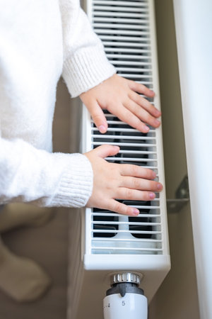Hands of little girl in warm sweater and socks standing by window and warming up from the heating...