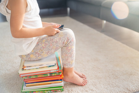 Little girl sits on a stack of children's fairy-tale books and watches cartoons on smartphone at sun...