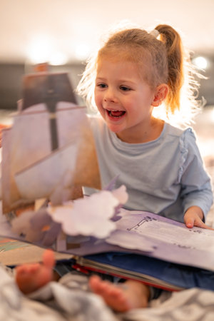 Little girl reading pop up book in home bed in Christmas environment with lights background. High...