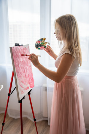 Young woman artist with palette and brush painting abstract pink picture on canvas near window. Art and creativity concept. High quality photoの写真素材