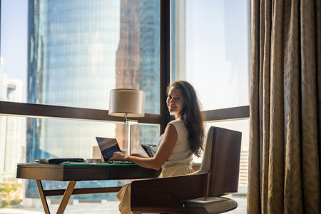 Stylish businesswoman freelancer working with laptop, she is sitting on the chair, panoramic view in window with city background. Low key photo. High quality photoの写真素材