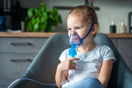Cute little girl are sitting and holding a nebulizer mask leaning against the face at home on sick leave, airway treatment concept. High quality photoの写真素材