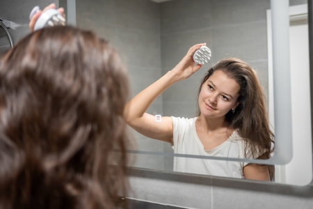 Young woman with dark hair doing self hair scalp massage with scalp massager or hair brush for hair growth stimulating at home bathroom. Reflected view of the mirror. High quality photoの写真素材