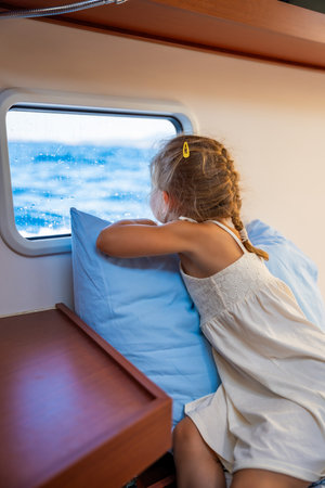 Blond little girl sits in the cabin of a yacht and looks out the window during a sea crossing. Yacht life concept. High quality photoの写真素材
