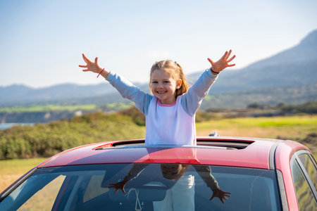 Little girl joyfully sticks out of car sunroof with raised hands against the backdrop of mountains in Northern Cyprus. The image symbolizes freedom, adventure, and the excitement of travel with child.の写真素材