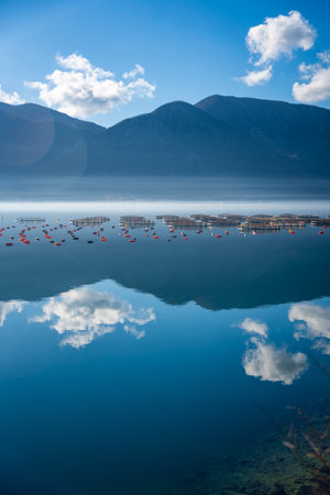Mountains with clouds and their reflection in the still waters of the sea, separated by a strip of fog in winter morning.の写真素材