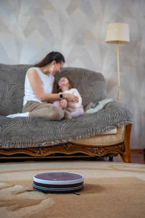 Robot vacuum cleaning the floor while a mother and daughter relax together on the couch in the background. Concept of smart home technology freeing time for family connection. High quality photoの写真素材