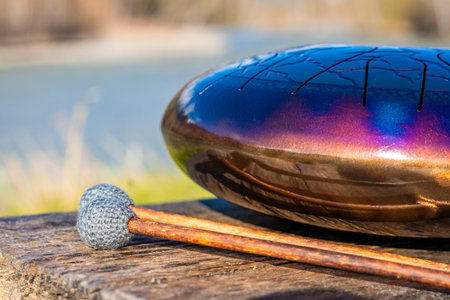 Steel tongue drum and mallets resting on a wooden bench with mountains, river, and forest in the...