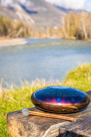 Steel tongue drum and mallets resting on a wooden bench with mountains, river, and forest in the...