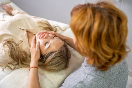 Female practitioner performs facial lifting technique on a young woman lying on a couch. Concept of energy healing, face therapy and alternative wellness practice. High quality photoの写真素材