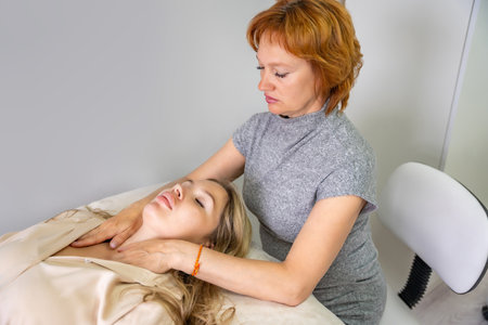 Female practitioner performs facial lifting technique on a young woman lying on a couch. Concept of energy healing, face therapy and alternative wellness practice. High quality photoの写真素材