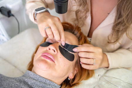 Beautician applying black eye patches before eyelash extension procedure with ultraviolet lamp....