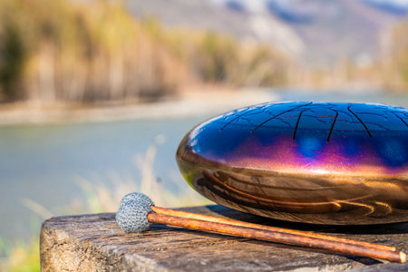 Steel tongue drum and mallets resting on a wooden bench with mountains, river, and forest in the...