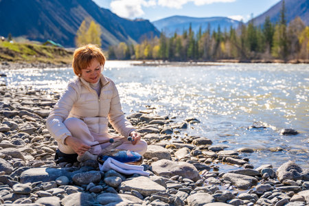Woman playing a steel tongue drum near a mountain river in Altai, surrounded by wild nature. Concept of spiritual retreat, sound healing, and ancient connection to earth. High quality photoの写真素材