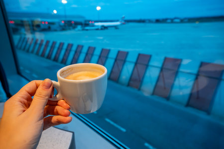 Hand holding coffee cup with night view of airport and planes in background. Concept of late-night flights, travel rituals, and quiet moments before departure. High quality photoの写真素材