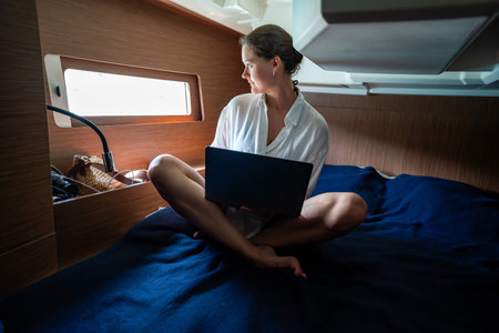 Young woman working with laptop and smartphone in the cabin of a sailing yacht near Croatia. Remote...