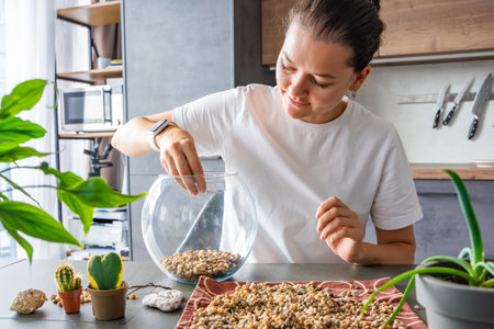 Young woman pouring stones into a glass container while preparing a terrarium. Concept of mindful crafting, eco-friendly lifestyle and home plant design. High quality photoの写真素材