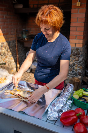 Woman seasoning fish and wrapping it in foil for outdoor grilling in her backyard. Homemade cooking, natural ingredients, and traditional barbecue preparation. High quality photoの写真素材
