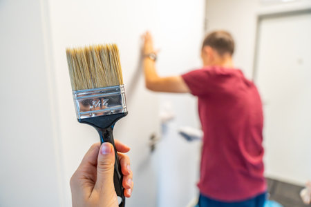 Close-up of a hand holding a paintbrush with a man painting the wall in the background. Concept of manual work, home renovation and attention to detail during interior improvement. High quality photoの写真素材
