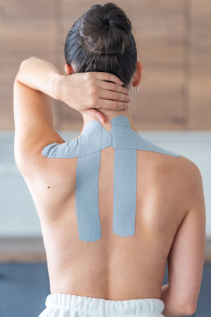 Young woman with kinesiology tape applied to her neck and shoulder holding her neck with her hand. Physical therapy and muscle recovery through modern rehabilitative care. High quality photoの写真素材
