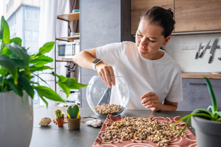 Young woman pouring stones into a glass container while preparing a terrarium. Concept of mindful crafting, eco-friendly lifestyle and home plant design. High quality photoの写真素材