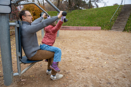 Man and child sitting and working out together with sports machine in the park. Shared workout...