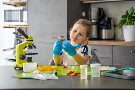 Girl wearing protective gloves examining grown crystal at table with microscope and laboratory...