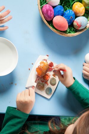 Overhead view of the hands of a child seated at a table painting colourful Easter eggs with a basket full of completed ones alongsideの写真素材