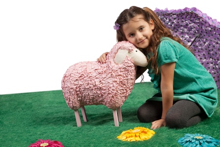 Cute pretty little girl sitting on the floor on an artificial meadow cudddling a large pink woolly toy sheep with a lovely smile on her faceの写真素材
