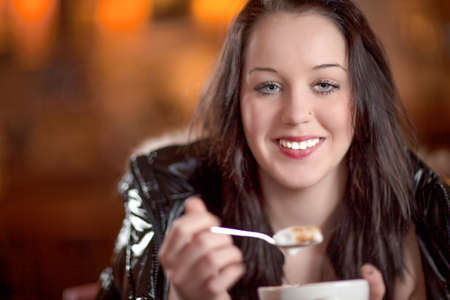 Smiling woman eating froth off her cappuccino coffee as she sits in a restaurant with a bokeh of orange lights behind herの写真素材