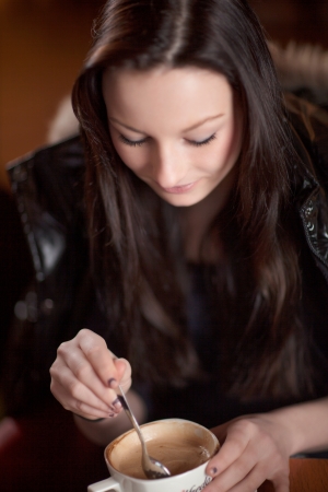 Attractive young woman enjoying a large cup of frothy cappuccino coffee in a restaurant stirring it with a spoonの写真素材