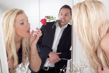 Reflection in the mirror of a nonchalant romantic man with a red rose between his teeth watching a beautiful blond woman titivating in the mirror looking up with a surprised lookの写真素材