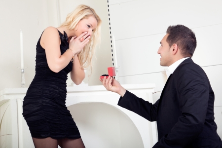 Handsome young man down on bended knee proposing marriage to a beautiful blond woman who is completely overwhelmed with surprise and joyの写真素材