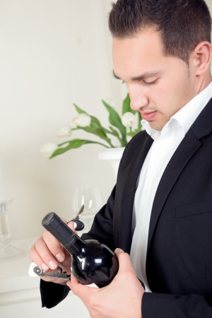 Stylish handsome young man in evening wear standing opening a bottle of red wine that he is holding in his handの写真素材