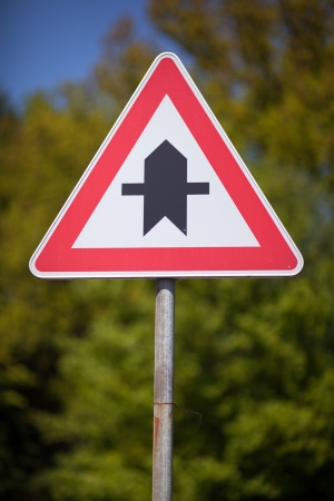 Triangular traffic sign indicating a crossroads on a main thoroughfare mounted on a roadside pole against greeneryの写真素材