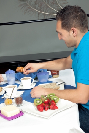 Young man having breakfast seated at a table in a restaurant with a variety of dishes stirring a cup of coffeeの写真素材