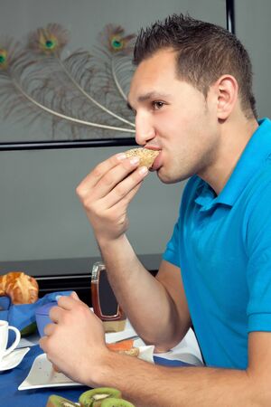 Young man eating breakfast biting into a fresh roll while seated at a table set with fresh fruit and coffeeの写真素材