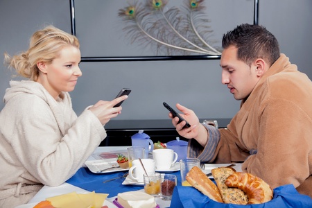 Candid portrait of a young couple in bathrobes eating breakfast in the early morning and each consulting their mobile phones for messagesの写真素材