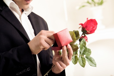 Closeup torso image of the hands of a young man about to propose holding a single long stemmed red rose and checking to make sure the ring is inside its boxの写真素材