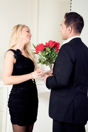 Romantic couple standing in evening attire in front of a fireplace with a vase of red roses talking and looking into each others eyesの写真素材