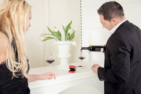 Man pouring red wine with a open ring box between the glasses on top of the mantelpiece as he is watched by his girlfriend or wifeの写真素材